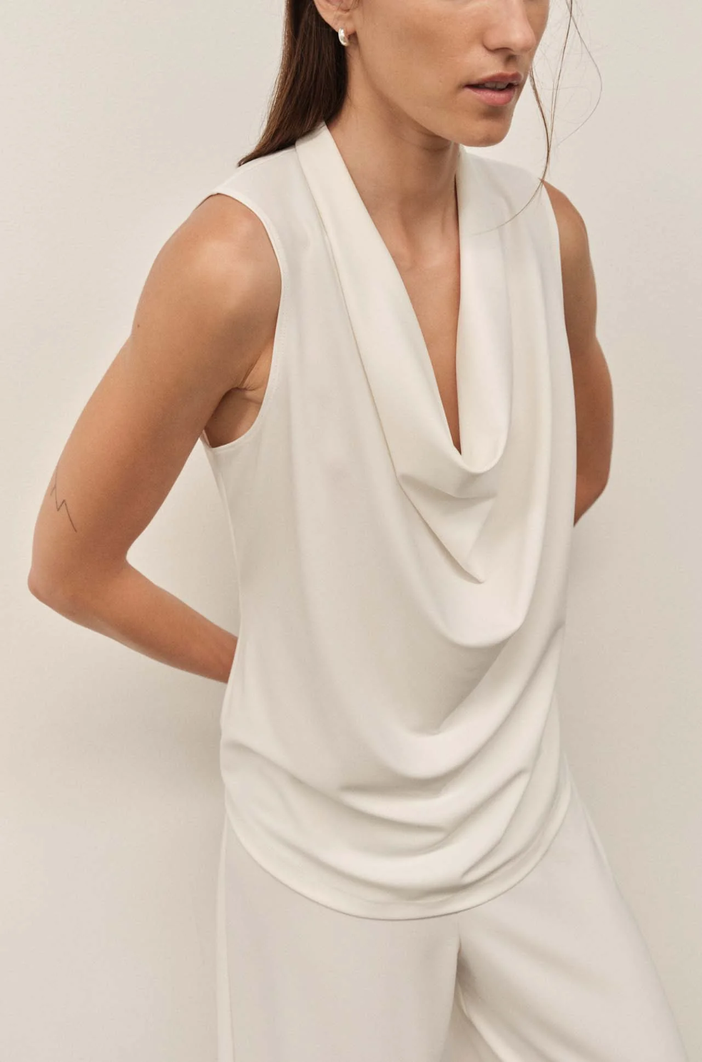 Woman wearing a white sleeveless top with a draped design on a plain background