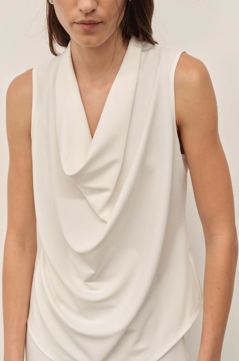 Woman wearing a white sleeveless top with a draped design on a plain background