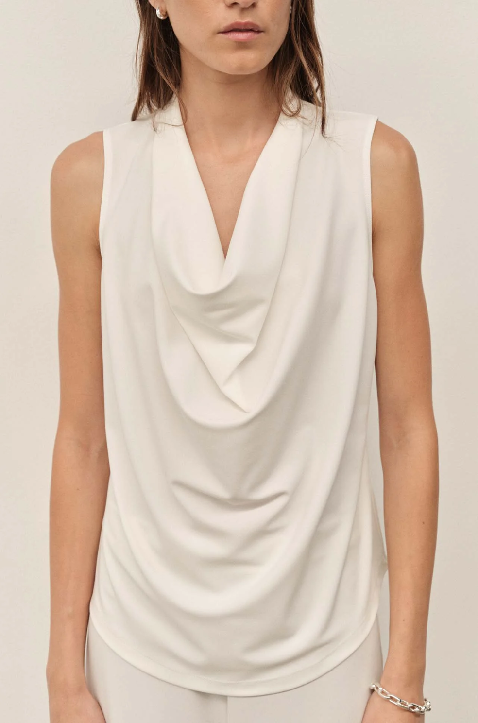 Woman wearing a white sleeveless top with a draped neckline on a plain background