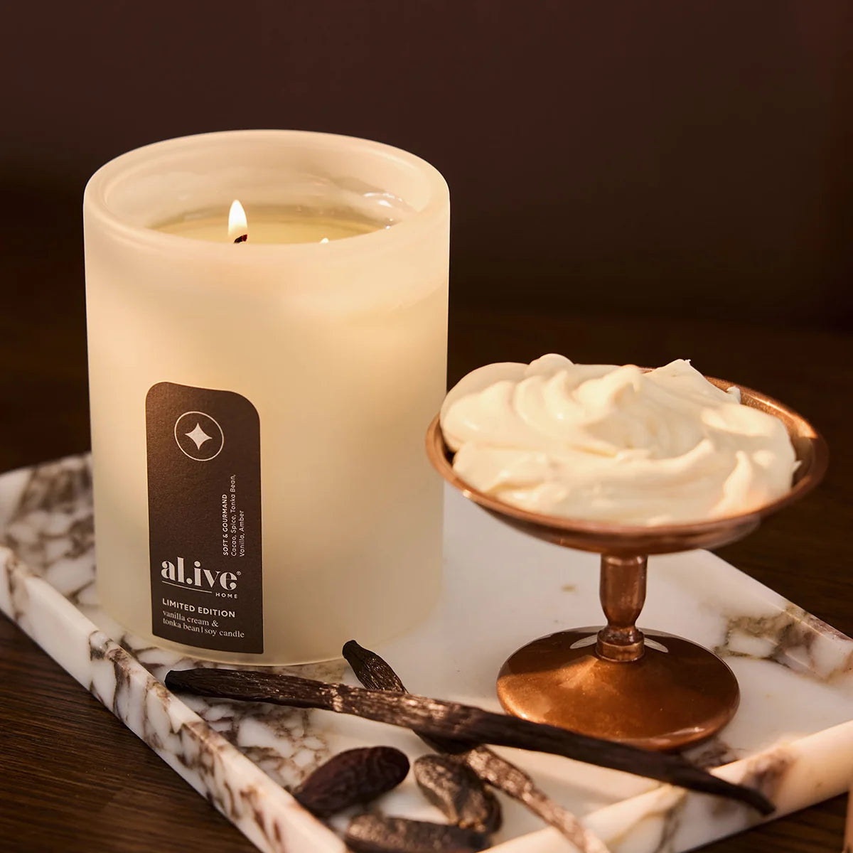 al.ive body Vanilla Cream & Tonka Bean Soy Candle lit on a marble tray with vanilla pods