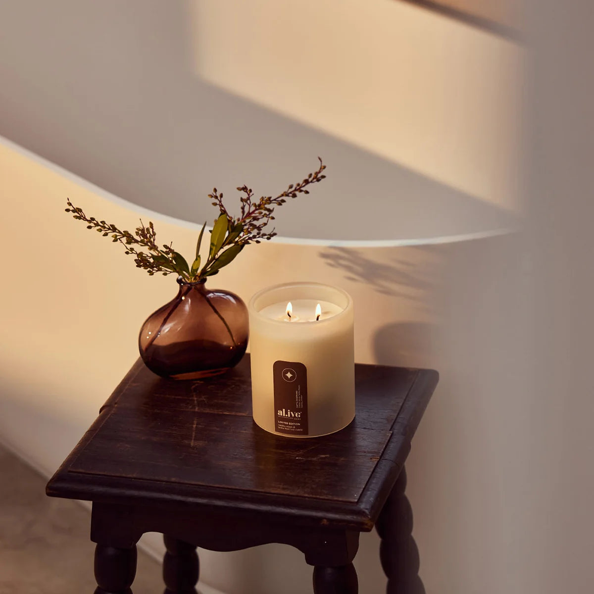 al.ive body Vanilla Cream & Tonka Bean Soy Candle lit on a timber side table with dried flowers
